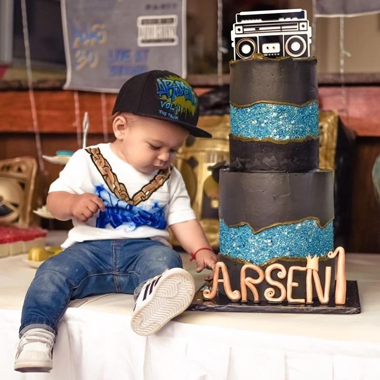 One year old baby boy sitting by two-tier boom box themed birthday cake with blue and gold accents, by Nadiya's Cupcakes
