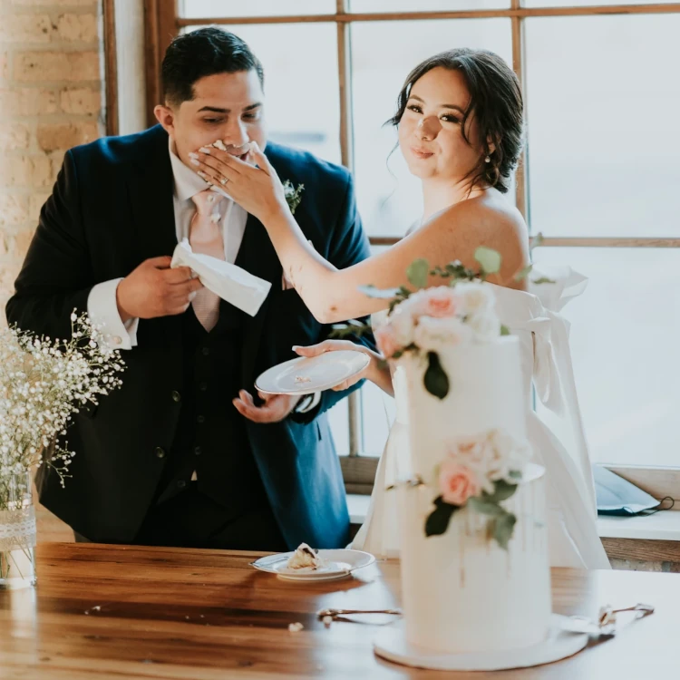 Wedding couple feeding each other near two-tier wedding cake with drips and fresh flower details, Carpentersville, by Nadiya's Cupcakes