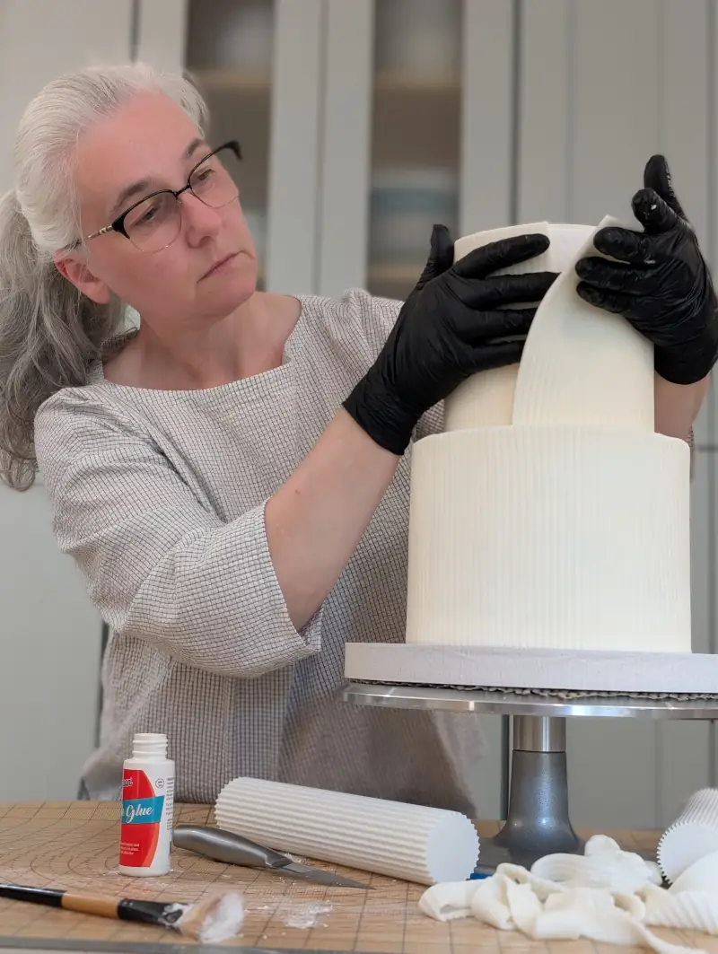 Nadiya wrapping textured gum paste onto two-tier cake, by Nadiya's Cupcakes