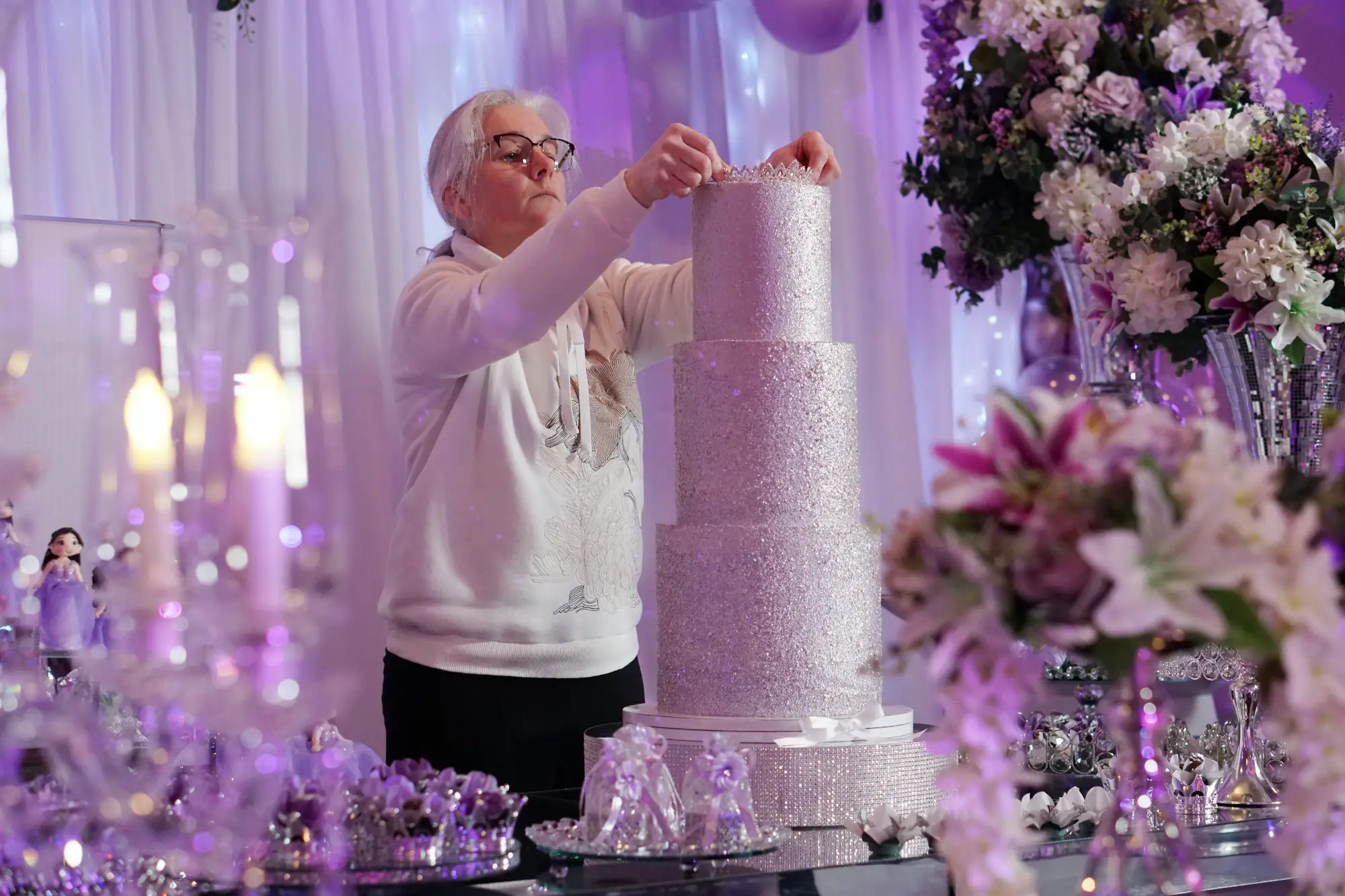 Nadiya putting a silver tiara on a three-tier pastel purple birthday cake decorated with silver glitter, by Nadiya's Cupcakes