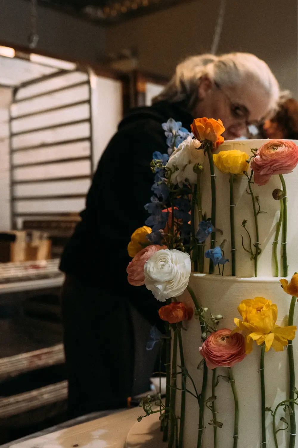 Nadiya working in the background preparing with a two-tier wedding cake decorated in fresh garden flowers in front of her, by Nadiya's Cupcakes