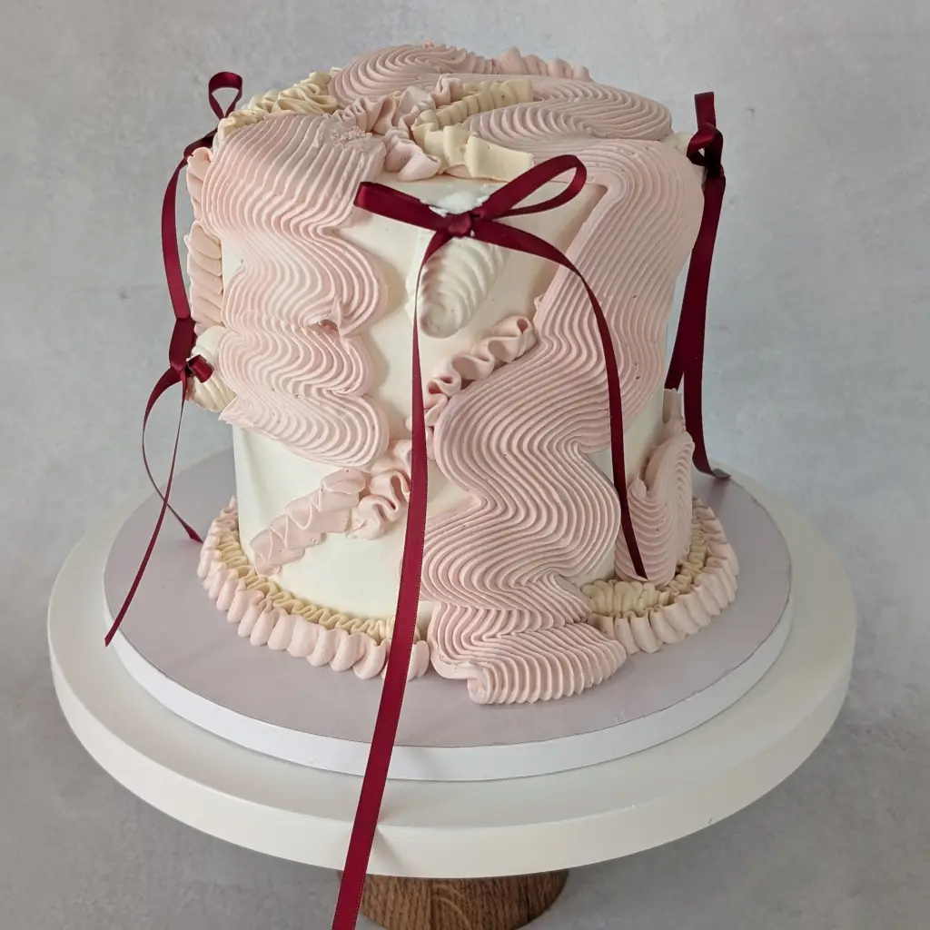 Top angle of a buttercream cake piped in various flowy piping styles with small maroon bows on a cake stand, by Nadiya's Cupcakes