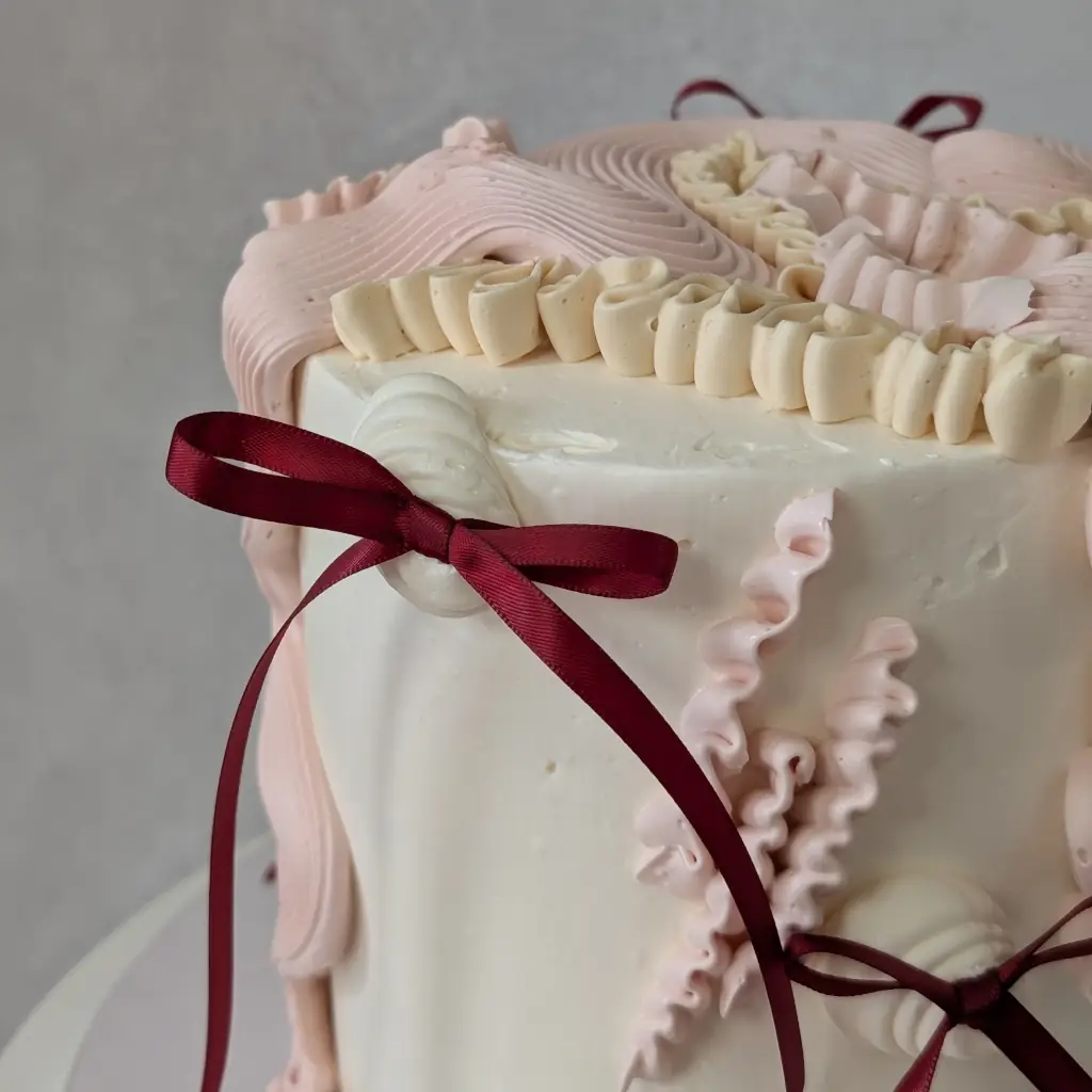 Top angle close-up of a buttercream cake piped in various flowy piping styles with small maroon bows on a cake stand, by Nadiya's Cupcakes