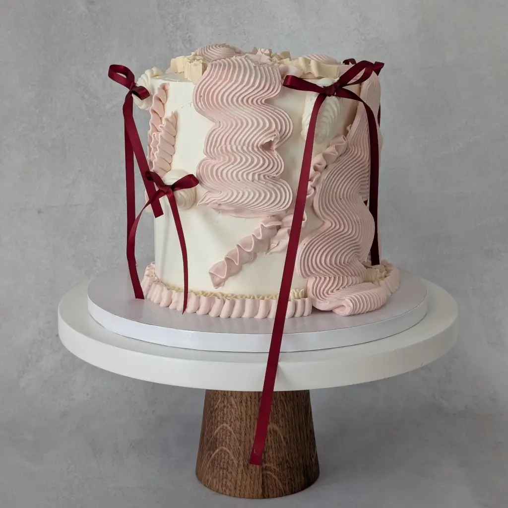 A buttercream cake piped in various flowy piping styles with small maroon bows on a cake stand, by Nadiya's Cupcakes