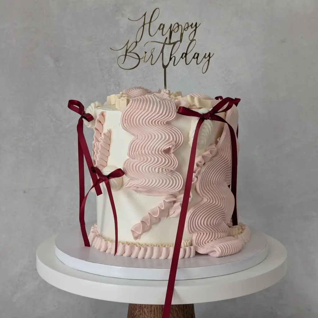 A buttercream cake piped in various flowy piping styles with small maroon bows and gold acrylic happy birthday cake topper, by Nadiya's Cupcakes