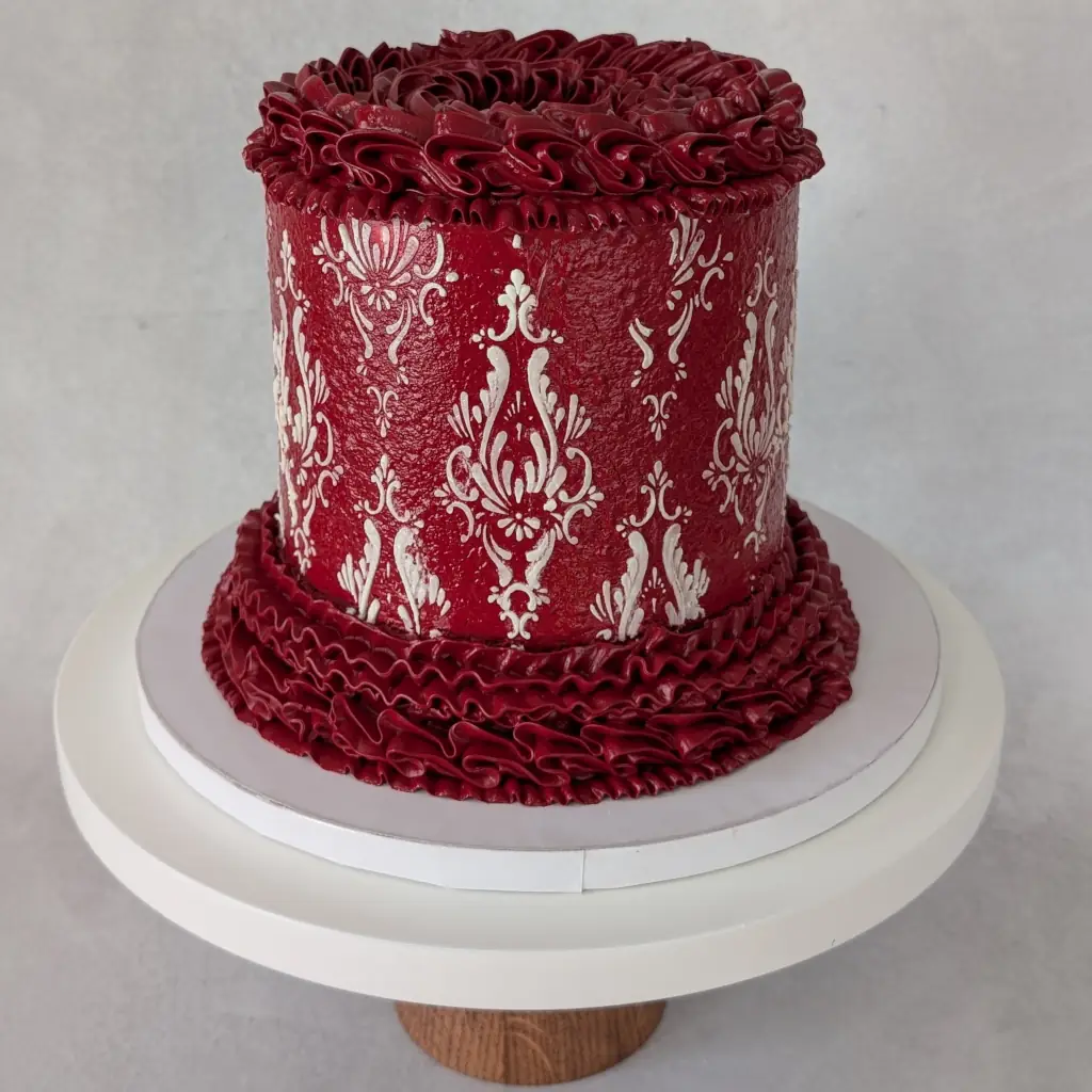 Angle view of a red buttercream piped cake with white buttercream stencils on a cake stand, by Nadiya's Cupcakes
