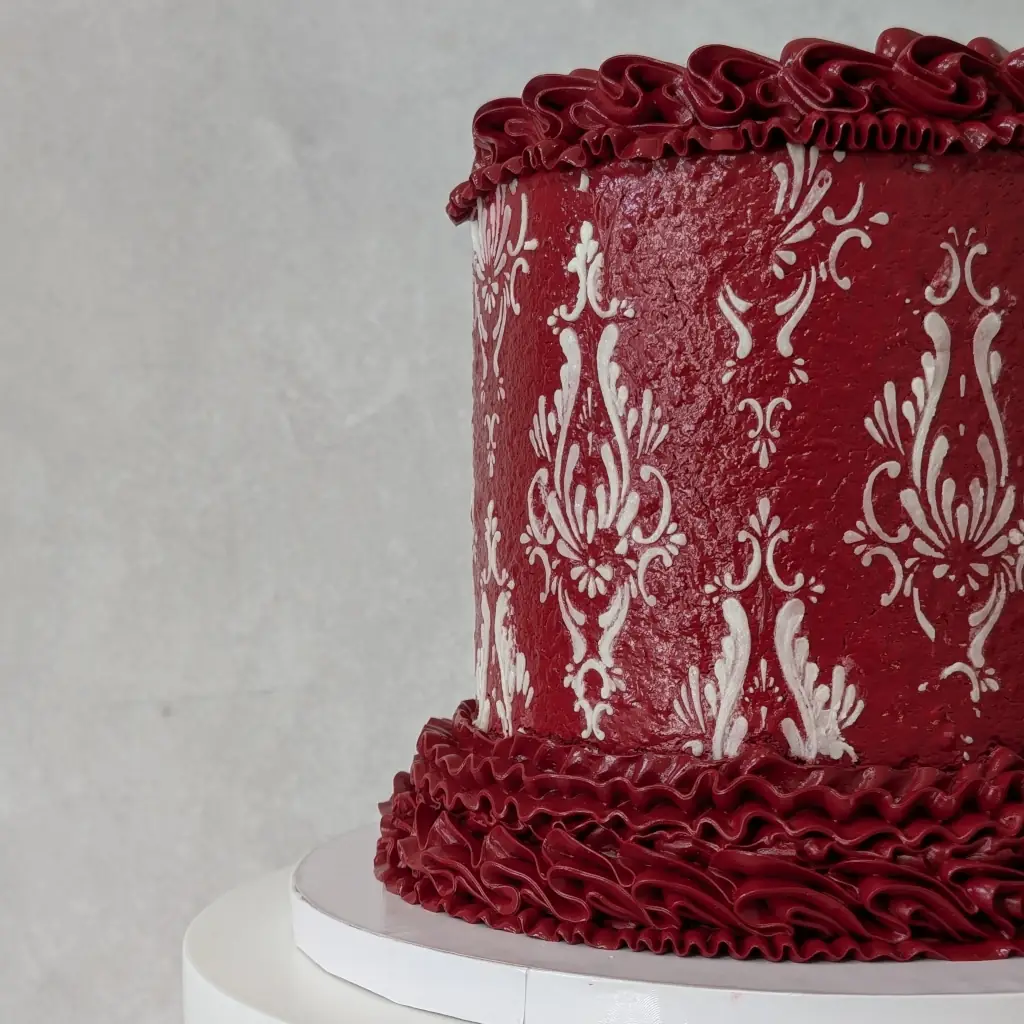 Close-up side view of red buttercream piped cake with white buttercream stencils on a cake stand, by Nadiya's Cupcakes