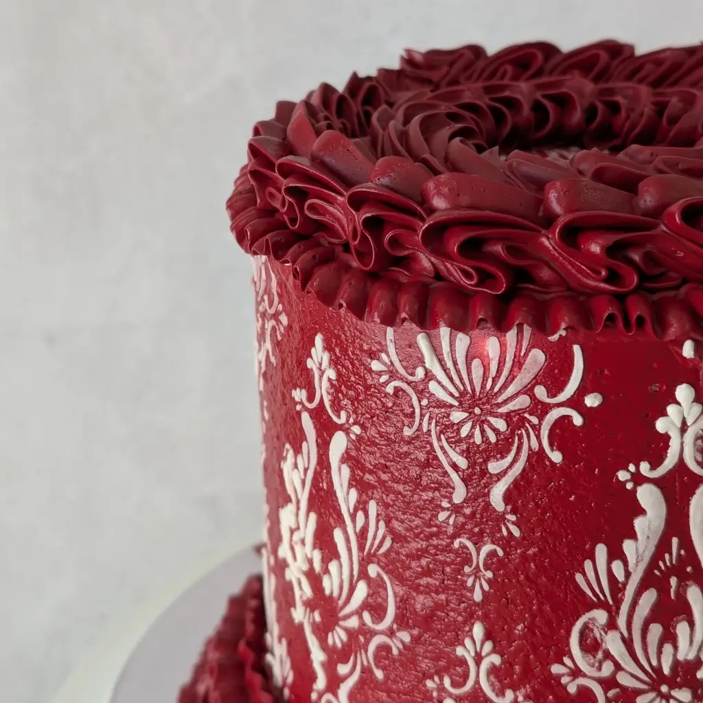 Close-up top view of red buttercream piped cake with white buttercream stencils on a cake stand, by Nadiya's Cupcakes